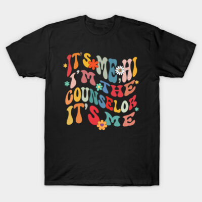 Black t-shirt with colorful, playful text design featuring the phrase "It's me, hi, I'm the counselor, it's me" and floral accents.
